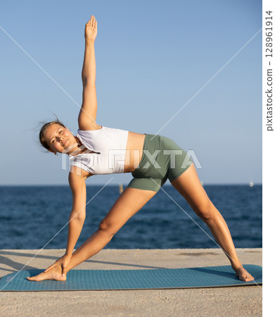Sportive girl practicing triangle pose of yoga on seashore 128961914