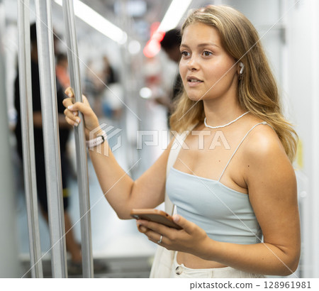 Young woman is standing in subway car with headphones and phone, getting to work Young woman is standing in subway car with headphones and phone, getting to work 128961981