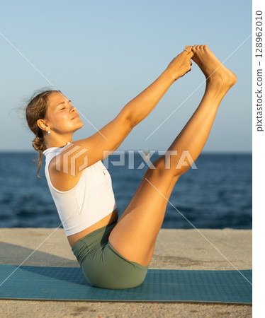 On windy day young slender woman does yoga near sea doing Ubhaya Padangushthasana 128962010