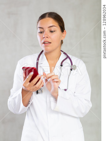 Female doctor in a white coat typing a message on mobile phone screen 128962034