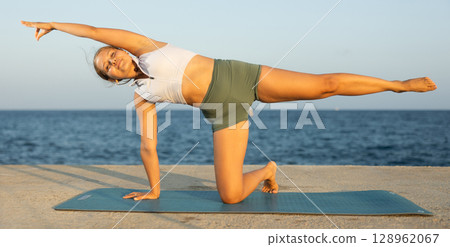 Sportive girl practicing kneeling half moon pose of yoga on seashore Sportive girl practicing kneeling half moon pose of yoga on seashore 128962067