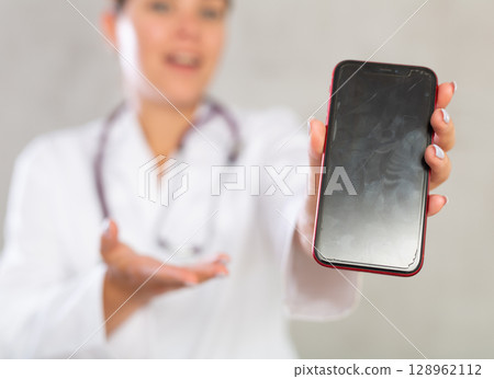 Female doctor in coat extending hand with phone and showing screen, selective focus on gadget. Online consultation concept 128962112