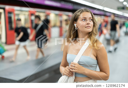 Girl with earphones is standing on subway platform, waiting for train to arrive 128962174
