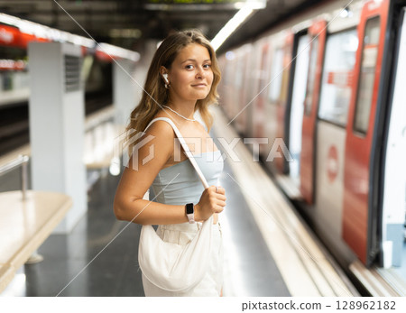 Girl with earphones is standing on subway platform, waiting for train to arrive Girl with earphones is standing on subway platform, waiting for train to arrive 128962182