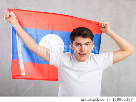 Happy young man holding flag of Laos against unicoloured background Happy young man holding flag of Laos against unicoloured background 128962397