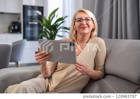 Blonde woman watching a show or movie on her digital tablet, enjoying quiet time in her living room. Technology and relaxation. Blonde woman watching a show or movie on her digital tablet, enjoying quiet time in her living room. Technology and relaxation. 128963787