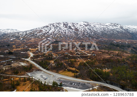 Magnificent Voringsfossen waterfall plunges into deep rugged canyon flanked by rocky cliffs. Winding road and sparse vegetation complete landscape 128963788