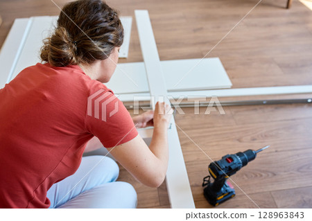 Woman sits on wooden floor and assembling furniture in new home, fastens parts of white bed frame. DIY home renovation and home organization after moving concept 128963843