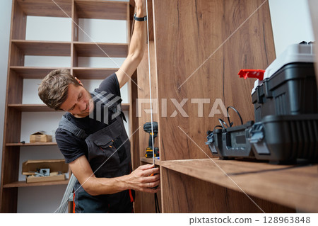 Close-up of man measuring wooden wardrobe with tape measure during furniture assembly, with tools and screws on shelf in background. Concept of home renovation and DIY furniture project Close-up of man measuring wooden wardrobe with tape measure during furniture assembly, with tools and screws on shelf in background. Concept of home renovation and DIY furniture project 128963848