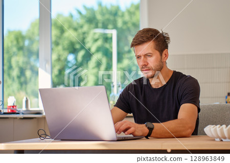 Man working on laptop at wooden table in bright kitchen. Freelancer working a home office. Concept of remote work and online business Man working on laptop at wooden table in bright kitchen. Freelancer working a home office. Concept of remote work and online business 128963849