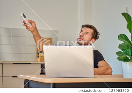 Man working on laptop at home and using remote control to adjust temperature on air conditioner. Person struggling with heat while working at home office Man working on laptop at home and using remote control to adjust temperature on air conditioner. Person struggling with heat while working at home office 128963850