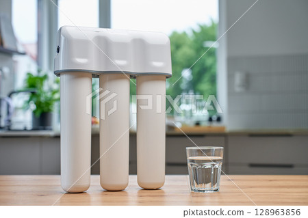 Glass of clean water on wooden kitchen table with water filtration system in background. Purified drinking water for healthy lifestyle 128963856