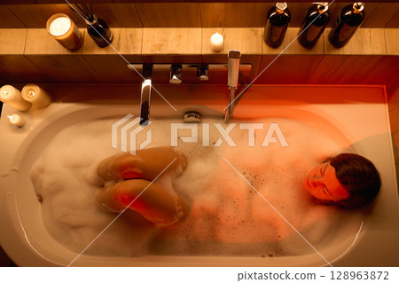Woman lies in bathtub filled with foam, top view. Woman relaxing in bathroom with candles and ambient lighting. Wellness, self care and home spa concept Woman lies in bathtub filled with foam, top view. Woman relaxing in bathroom with candles and ambient lighting. Wellness, self care and home spa concept 128963872