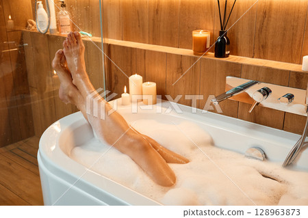 Female legs in bathtub filled with foam and warm lighting. Woman relaxing in bathroom with candles. Concept of wellness, relaxation, and self care 128963873