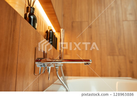 Modern chrome bathtub faucet with shower head mounted on wall with decorative candle and bottles. Contemporary bathroom interior 128963886