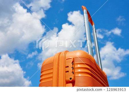 Bright orange suitcase with extended handle stands against blue sky with white clouds. Concept of travel, vacation and summer adventure 128963913
