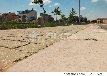 A deserted city street sidewalk from a lower angle, going into the distance along the embankment with a green fence and lighting poles. Public walking spaces in the hot summer period 128965269