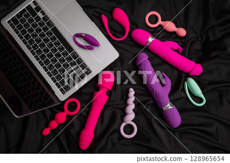 Collection of sex toys on a black sheet. Top view of various vibrators and a laptop.  128965654