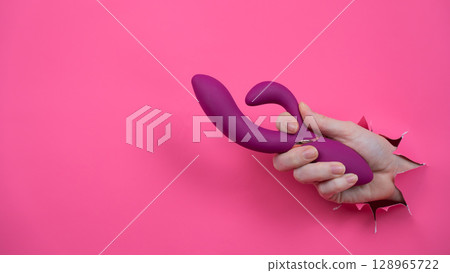 Female hand sticking out with raspberry sex toy from pink paper background. 128965722