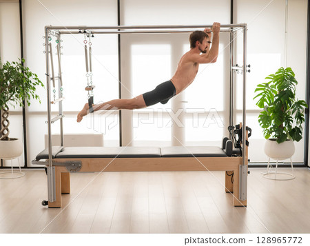 A man performs aerial exercises on a reformer machine. A man performs aerial exercises on a reformer machine. 128965772