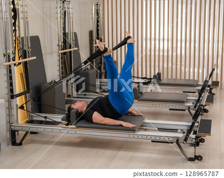 Overweight caucasian woman doing pilates exercises on a reformer. Overweight caucasian woman doing pilates exercises on a reformer. 128965787