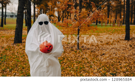 Woman in white sheet holding jack o'lantern against autumn forest background. Halloween costume. Woman in white sheet holding jack o'lantern against autumn forest background. Halloween costume. 128965884