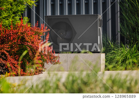 Modern outdoor heat pump unit near a metal wall, surrounded by green and red garden plants in a landscaped area 128965889