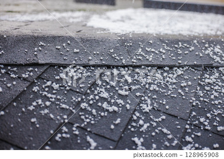 Close-up of small hailstones scattered on a black roof with shingles during a hailstorm. Close-up of small hailstones scattered on a black roof with shingles during a hailstorm. 128965908