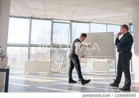 Two Caucasian men discussing work at a white board. Two Caucasian men discussing work at a white board. 128966000