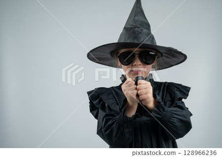 Portrait of a little Caucasian girl in a witch costume inflates a black balloon on a white background. Portrait of a little Caucasian girl in a witch costume inflates a black balloon on a white background. 128966236