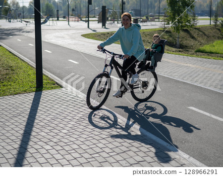 Caucasian woman riding bicycle with toddler son sitting behind her in child seat in park.  128966291