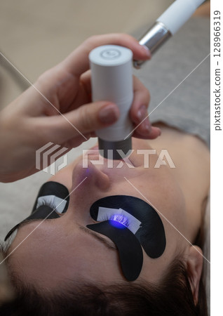 A master extends a woman's eyelashes using an LED lamp. Vertical photo.  128966319