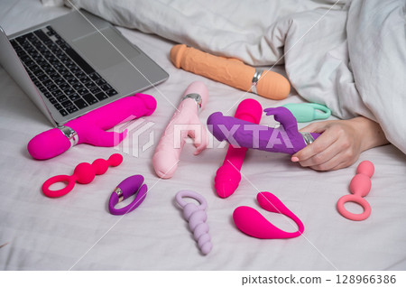 Woman holding sex toy. Various vibrators and laptop on bed.  128966386