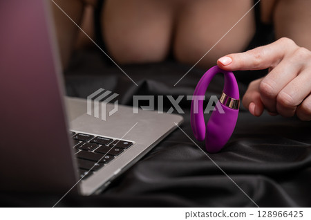 A woman is lying in bed holding a curved vibrator and looking at a laptop.  128966425