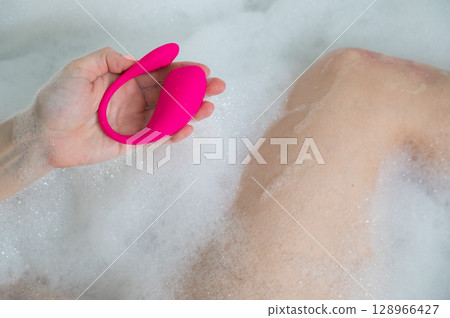 A woman lies in a bubble bath and holds a curved pink vibrator. 128966427
