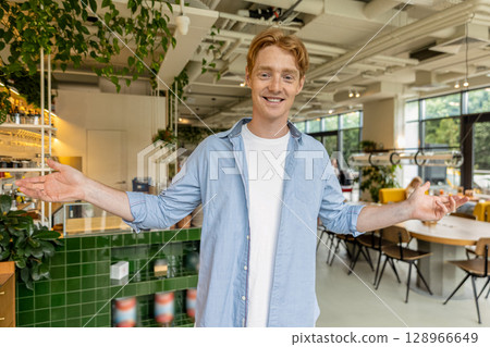 Ginger young guy in blue tshirt welcoming clients in a cafe Ginger young guy in blue tshirt welcoming clients in a cafe 128966649