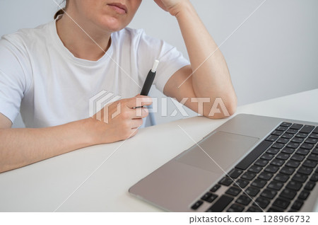A woman smokes an electronic cigarette while working on a laptop. Tobacco heating system. A woman smokes an electronic cigarette while working on a laptop. Tobacco heating system. 128966732