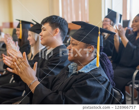 Young people in graduation gowns listen to a speech and applaud.  128966757