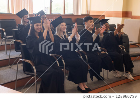 Young people in graduation gowns listen to a speech and applaud.  128966759