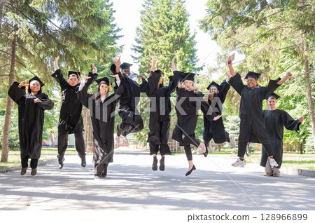 Happy multinational young people in graduation gowns jumping for joy outdoors.  128966899