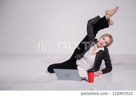 Barefoot ballerina dressed in a business suit poses for a laptop and drinks coffee. Flexible woman works at the computer 128967022
