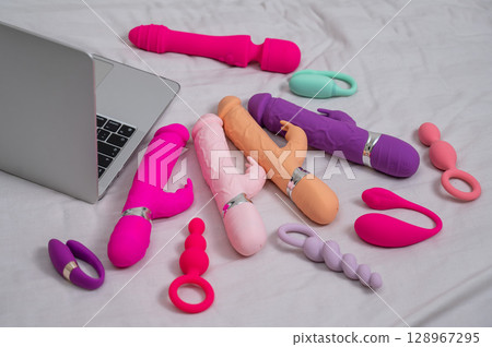 Collection of sex toys on a white sheet. Various vibrators and a laptop on the bed.  128967295