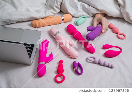 Woman holding sex toy. Various vibrators and laptop on bed.  128967296