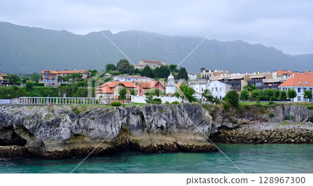Picturesque coastal town in Northern Spain, featuring traditional houses with red roofs perched atop dramatic cliffs overlooking a turquoise bay. Llane,s, Asturias 128967300