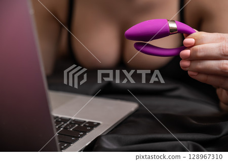 A woman is lying in bed holding a curved vibrator and looking at a laptop.  128967310