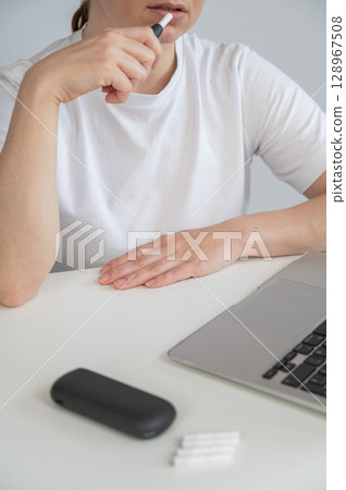 A woman smokes an electronic cigarette while working on a laptop. Tobacco heating system. A woman smokes an electronic cigarette while working on a laptop. Tobacco heating system. 128967508