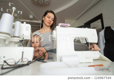 A young woman sews in the kitchen and holds a small child. Mom teaches her little son to sew on a sewing machine. Self-isolation homework 128967584