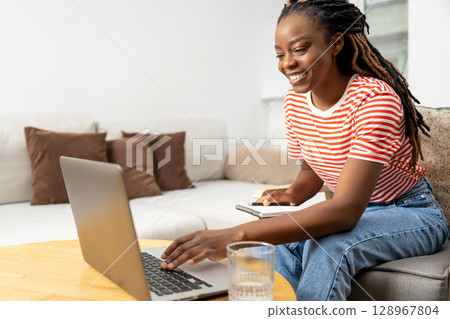 Smiling african american young woman sitting at the laptop and having a video call Smiling african american young woman sitting at the laptop and having a video call 128967804