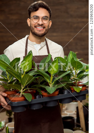 Smiling young man in eyeglasses holding flower pots and looking enjoyed 128969200
