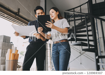 Construction manager and engineer dressed in orange work vests and hard helmets explore construction documentation on the building site near the steel frames 128969387